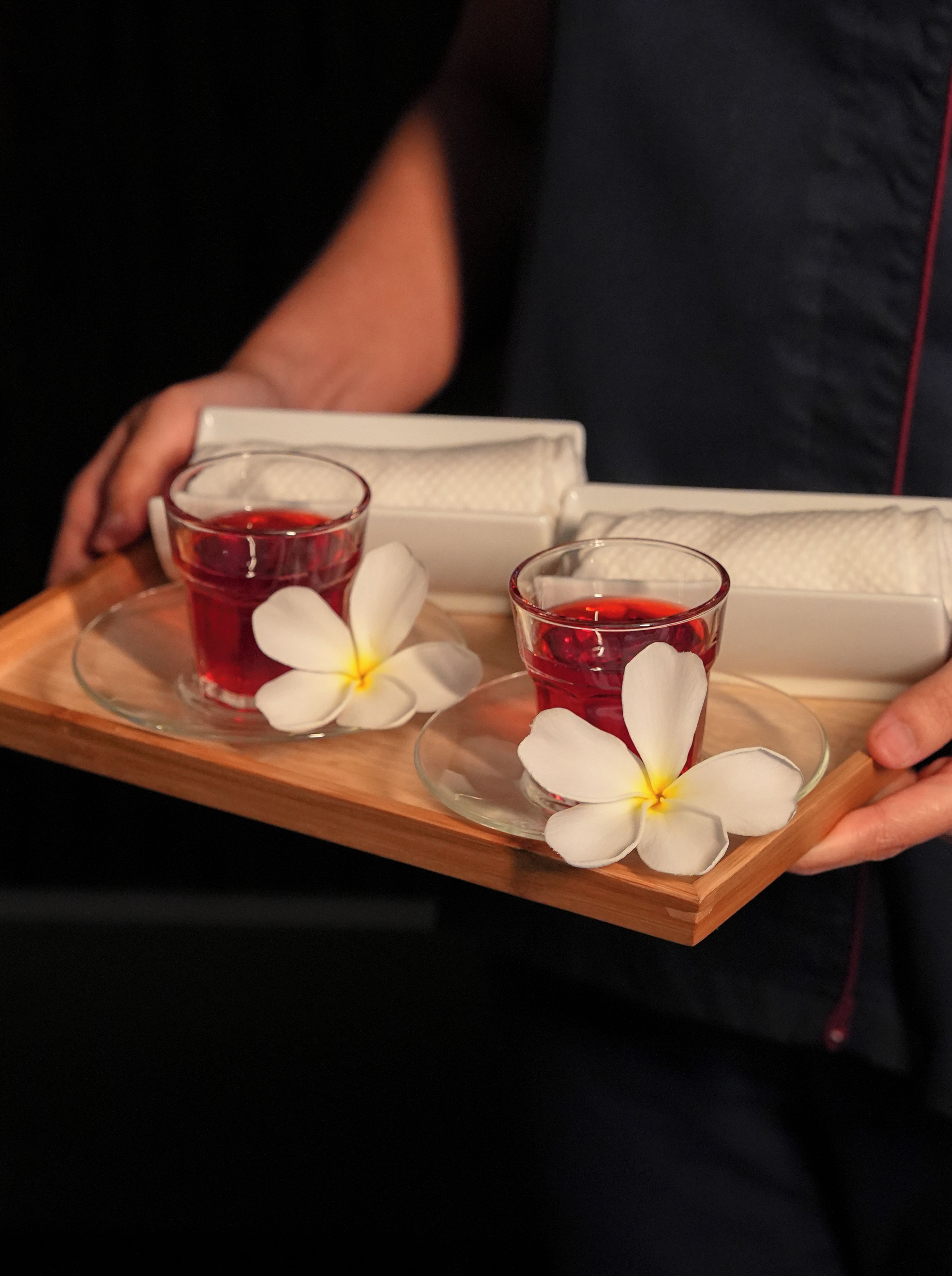 Una bandeja con dos vasos de bebida roja y dos flores blancas, sostenida por una persona.