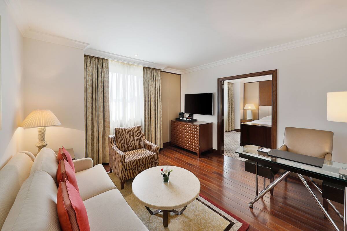 Habitación de hotel con sofá, sillón, mesa redonda de centro, televisión y escritorio con lámpara.