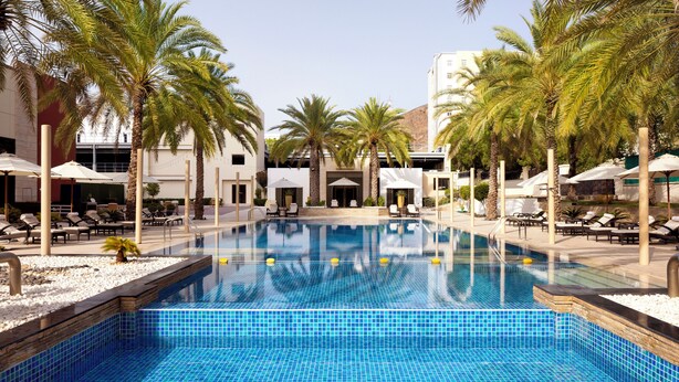 Una piscina amb gandules i ombrelles, envoltada de palmeres i un edifici amb disseny modern.