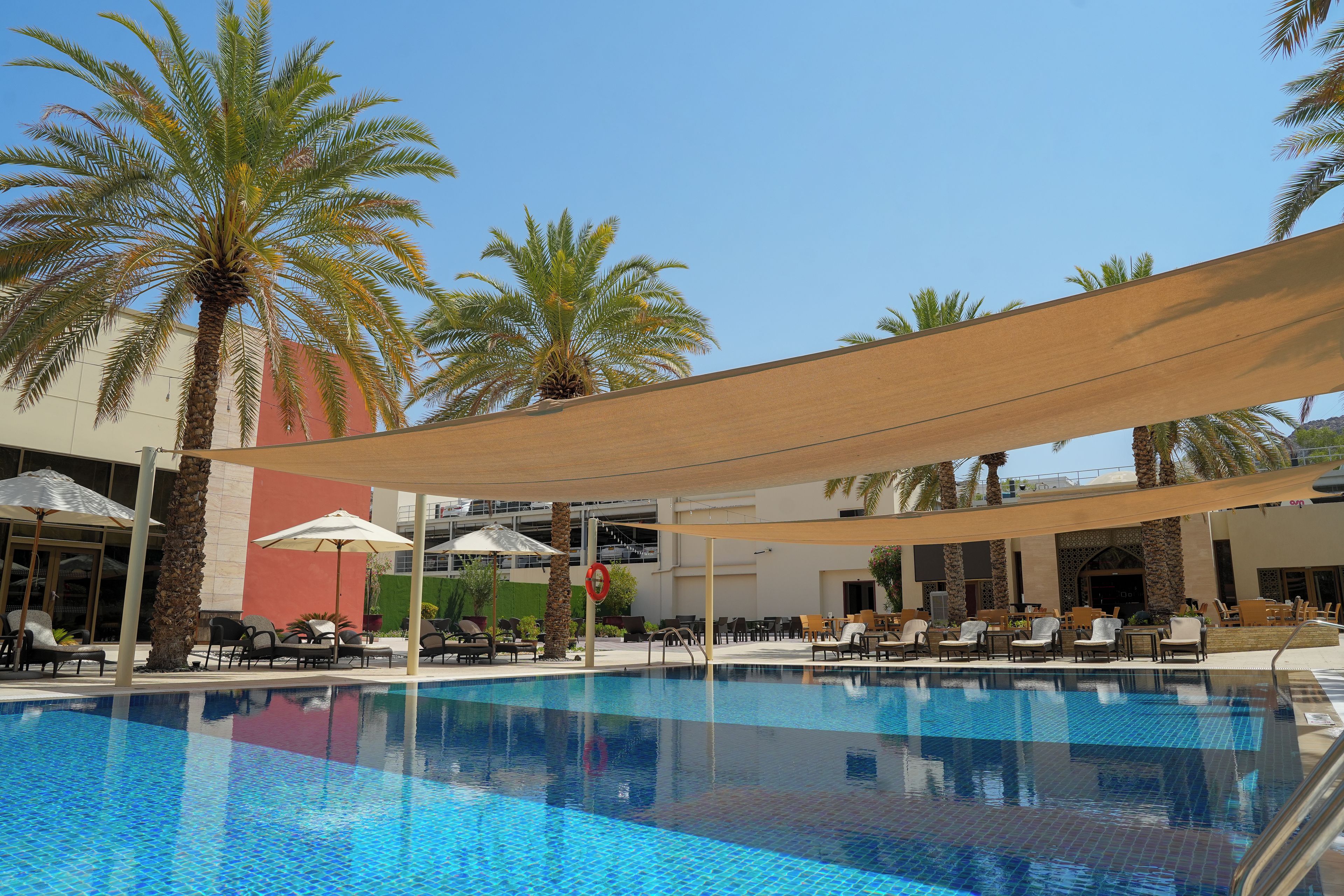 Ein Pool mit Liegestühlen und Sonnenschirmen, umgeben von Palmen und einem Gebäude mit roter Fassade.