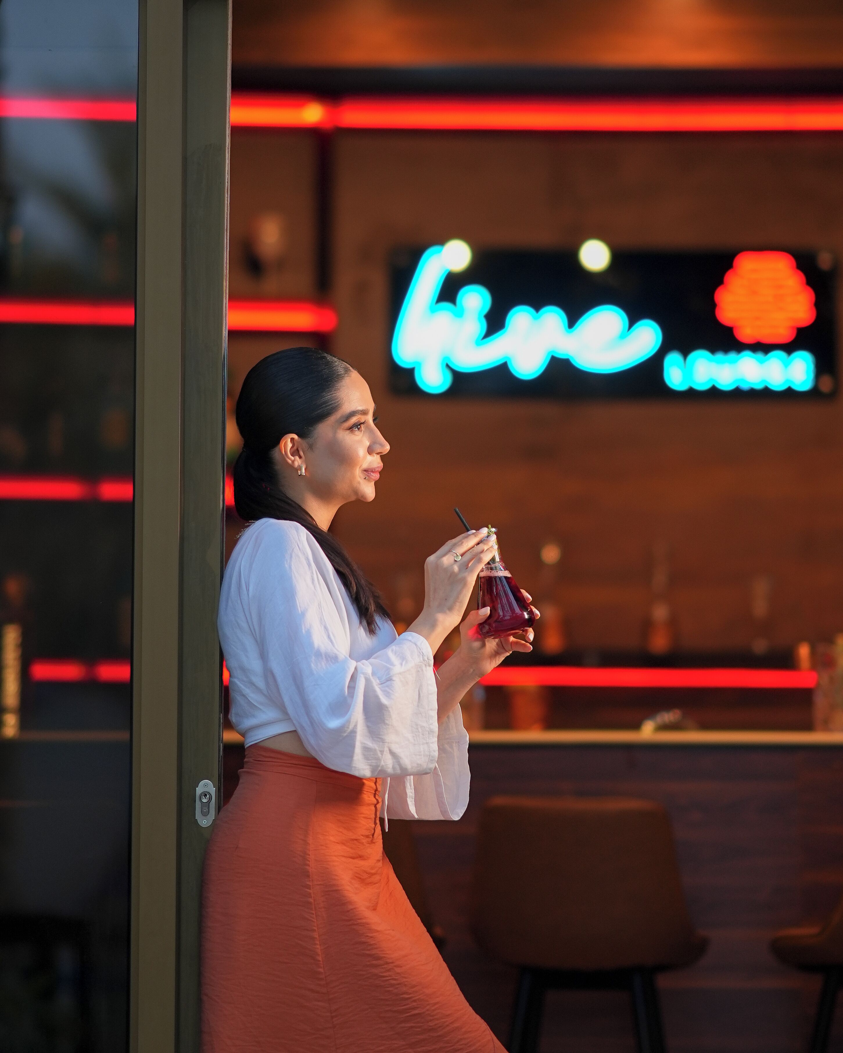 Una persona sosteniendo una bebida en un bar con un letrero de neón al fondo.