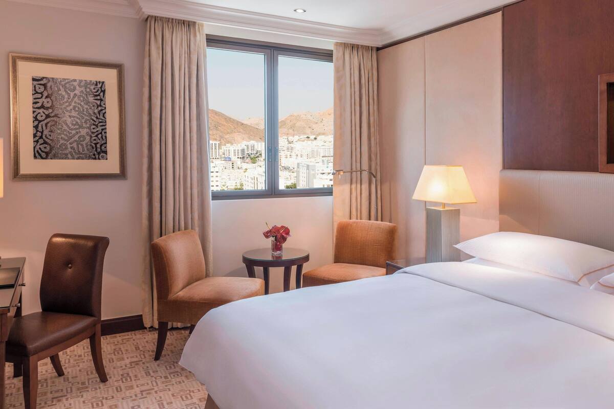 Habitación de hotel con una cama grande, dos sillones, una mesita con un jarrón, una lámpara y un cuadro en la pared.