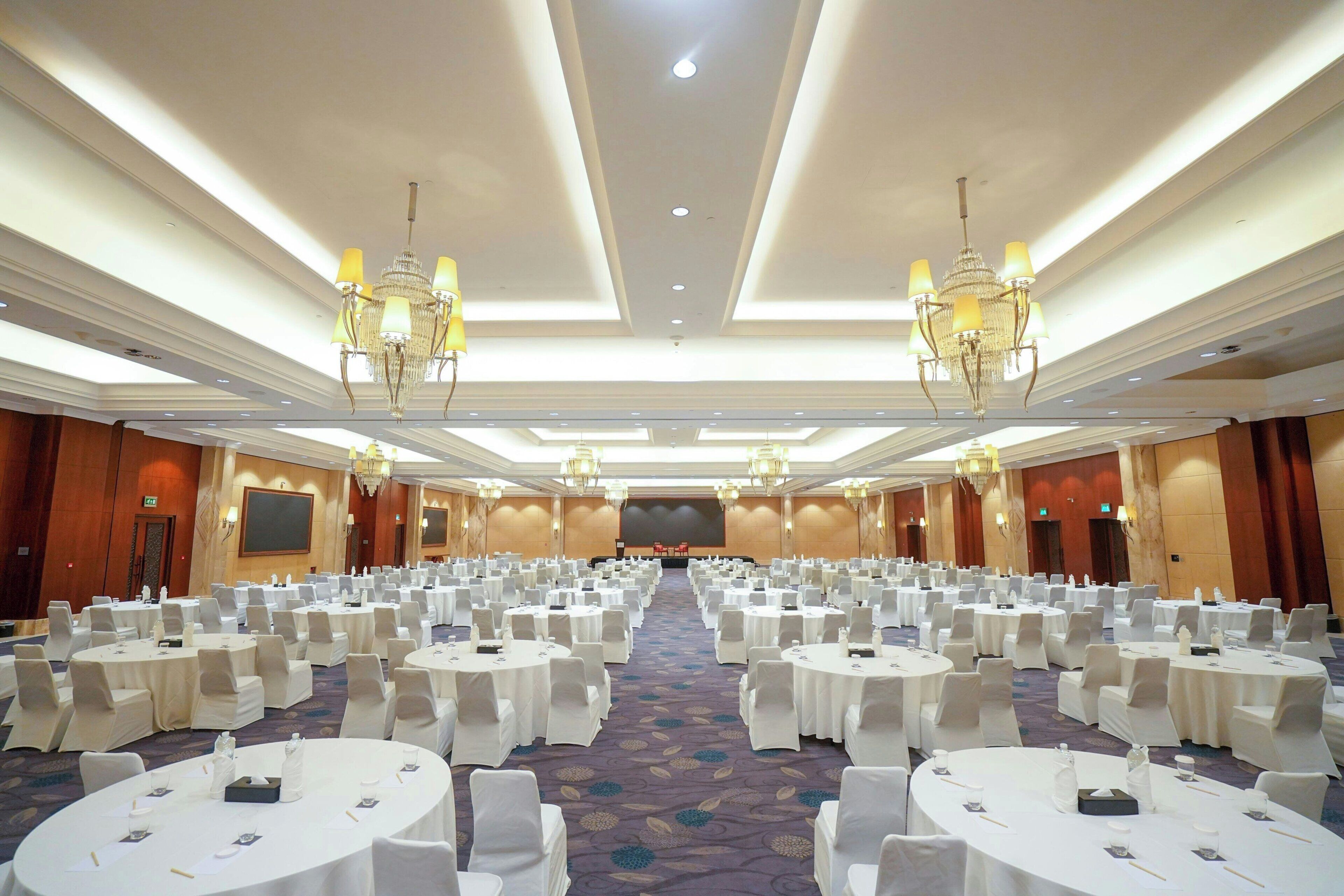 A large conference room with round tables, white chairs, chandeliers, and a carpeted floor.