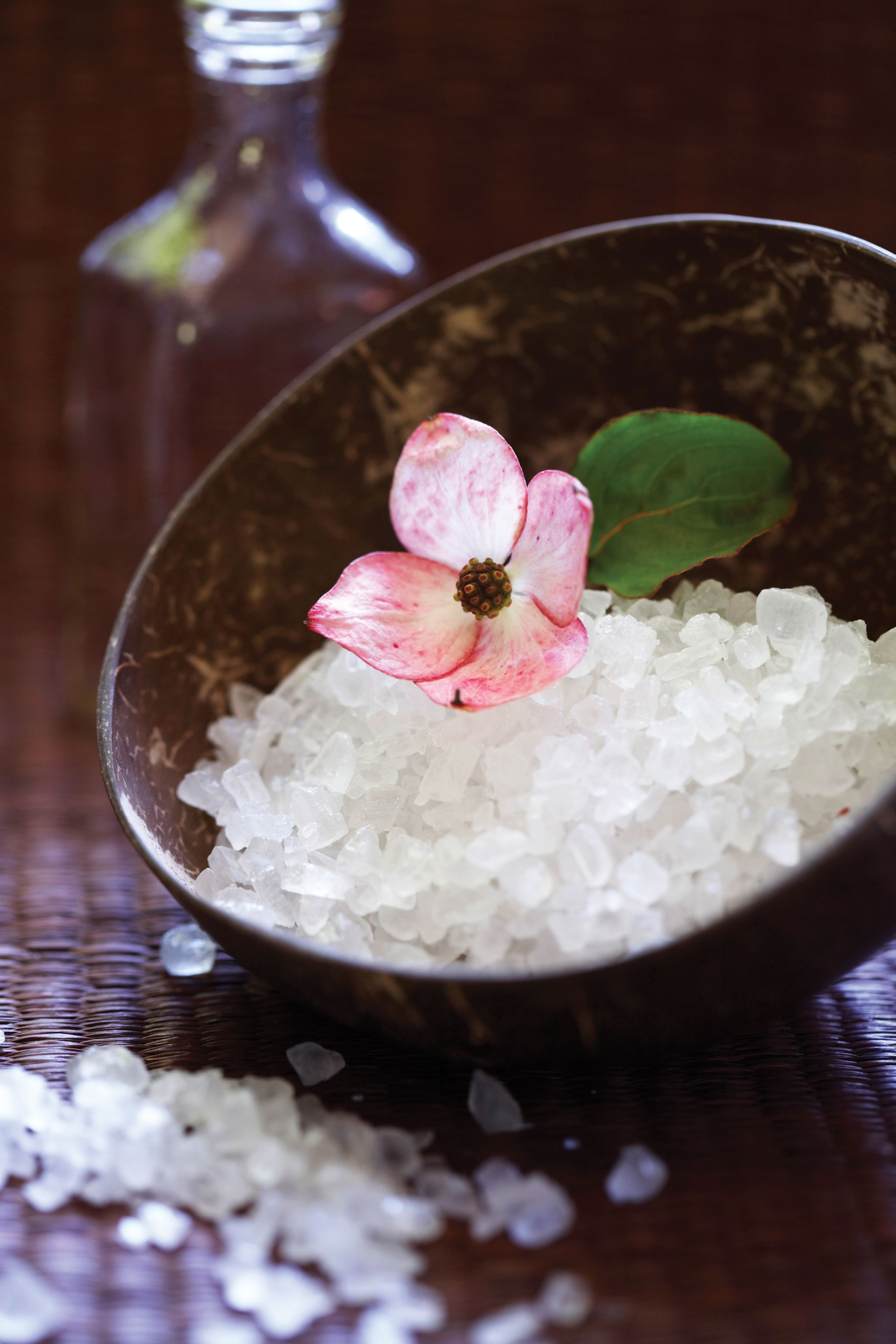 A bowl of white salt with a pink flower on top, placed on a wooden surface.
