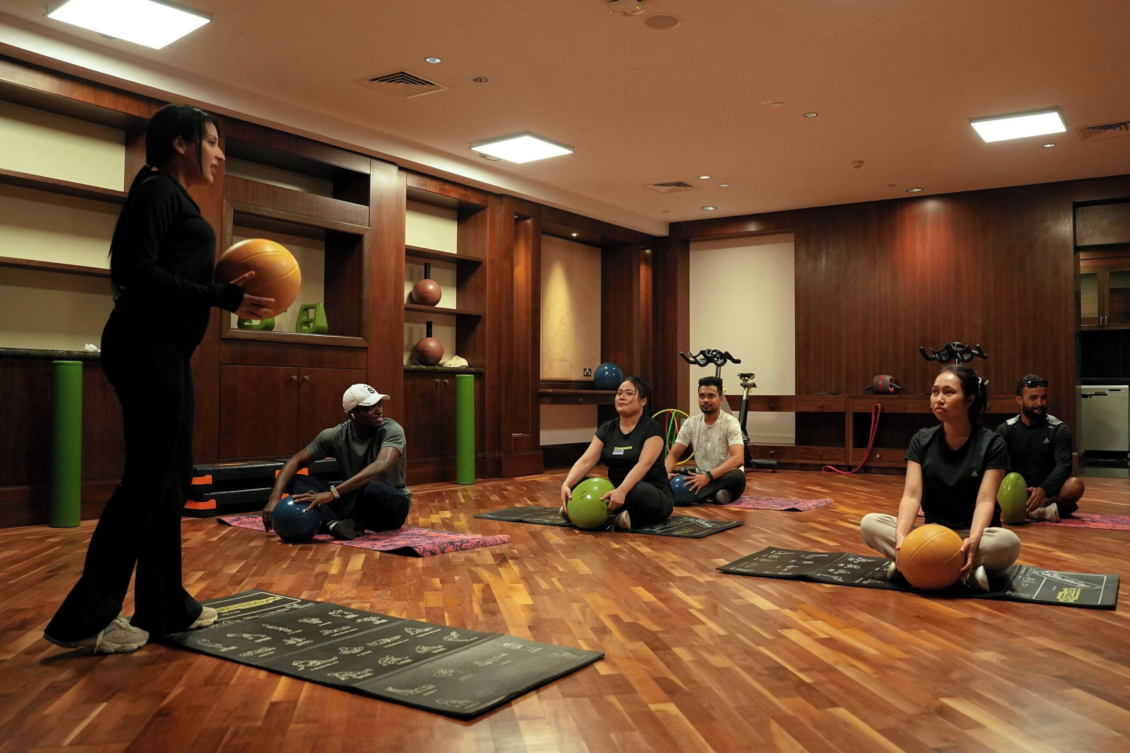 A group of people in a room with wooden floors and walls, engaged in a fitness class with exercise balls and mats.