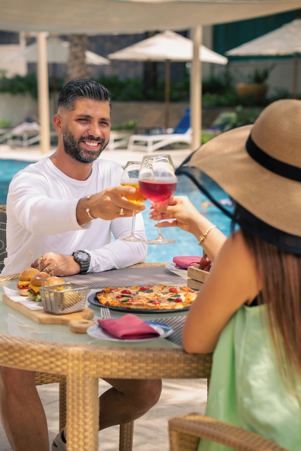 A dining table with food and drinks, a pool in the background, and outdoor umbrellas.