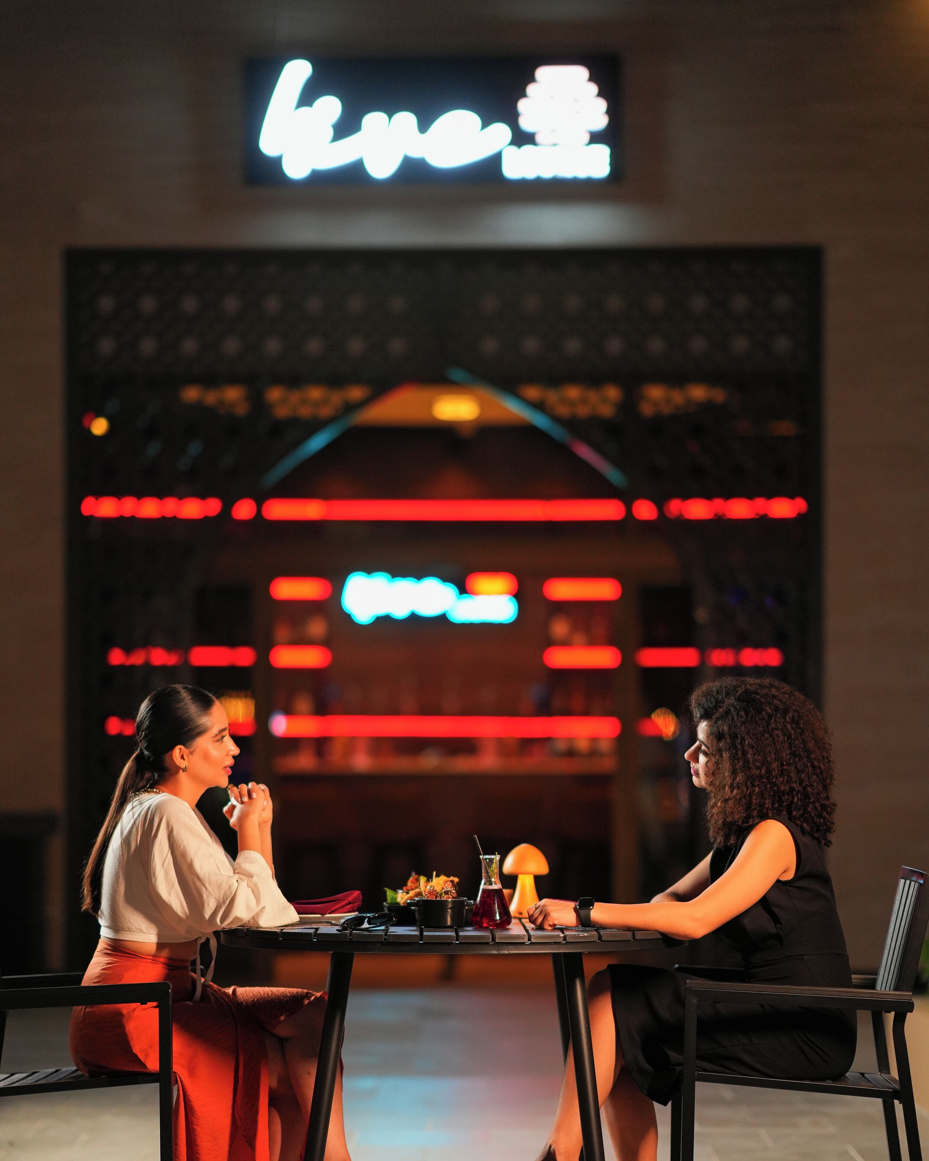 Two people seated at a table in a restaurant with a neon sign in the background.