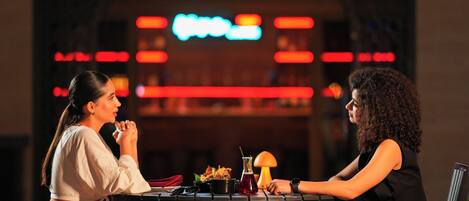 Two people seated at a table in a restaurant with a neon sign in the background.