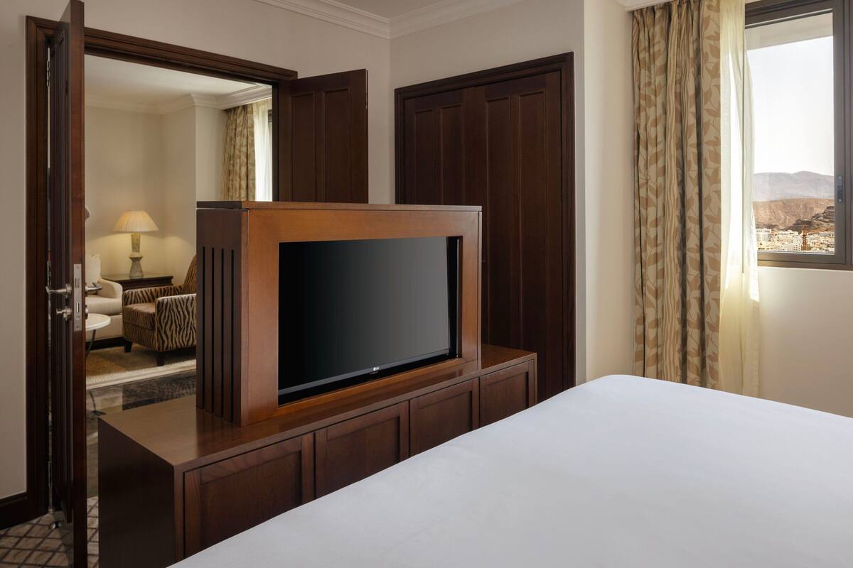 Habitación de hotel con un televisor de pantalla plana grande sobre un soporte de madera, una cama con ropa de cama blanca, un armario de madera y vistas a un paisaje montañoso a través de una ventana.