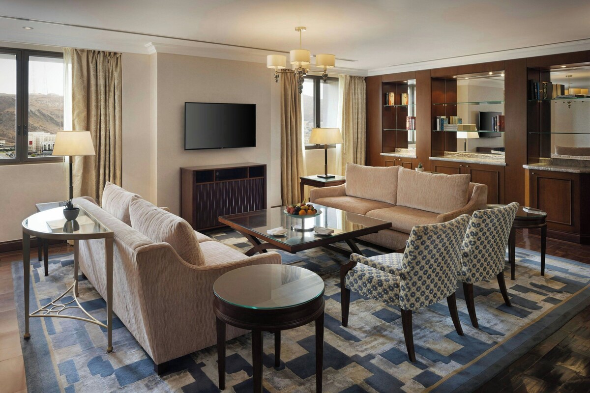 A hotel room with a sofa, armchairs, a coffee table, a TV, a fireplace, and a kitchen area.