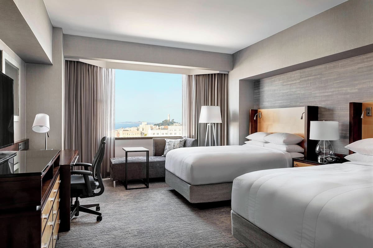 A hotel room with two beds, a desk, a chair, and a view of the city.