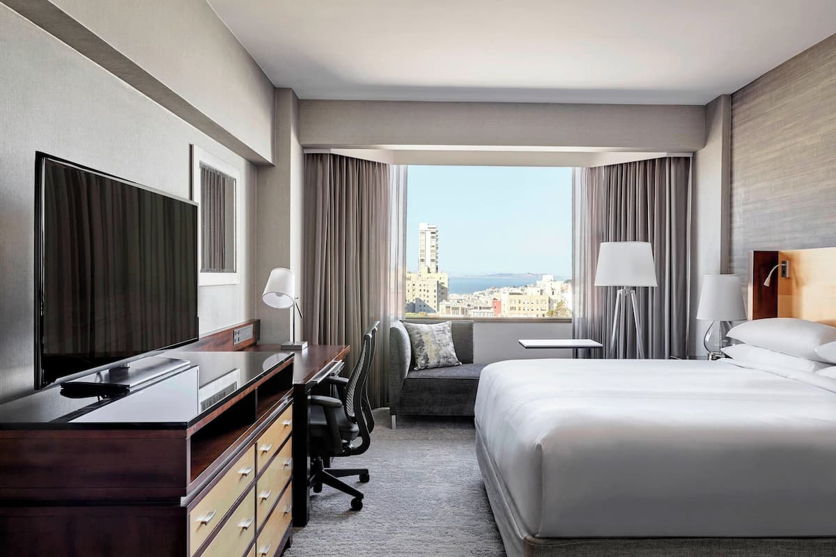 A hotel room with a bed, desk, chair, and a large window offering a city view.