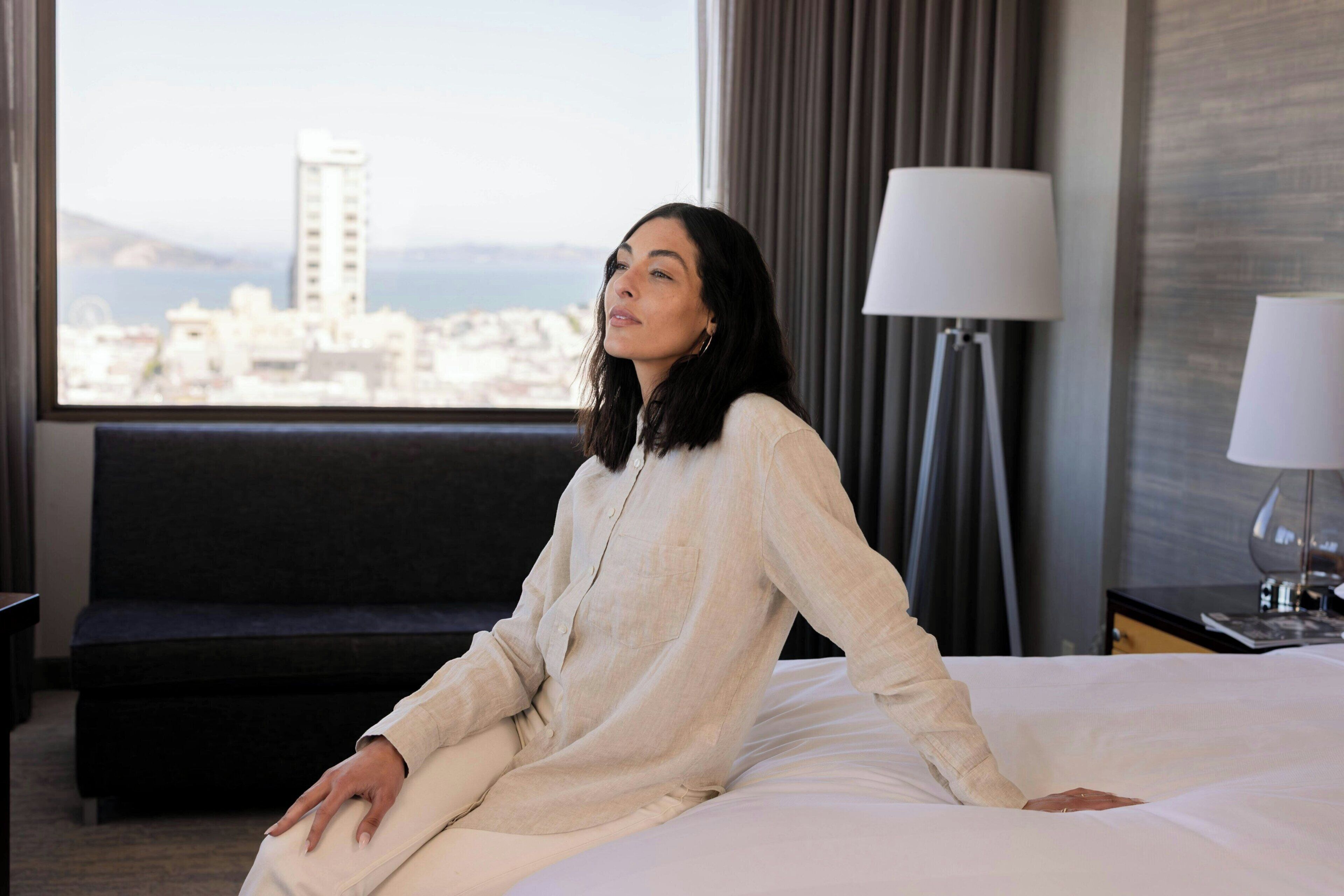 Una habitación de hotel con cama, ventana con vista a la ciudad, lámpara y sofá.