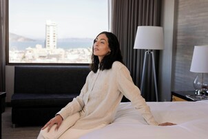Una habitación de hotel con cama, ventana con vista a la ciudad, lámpara y sofá.