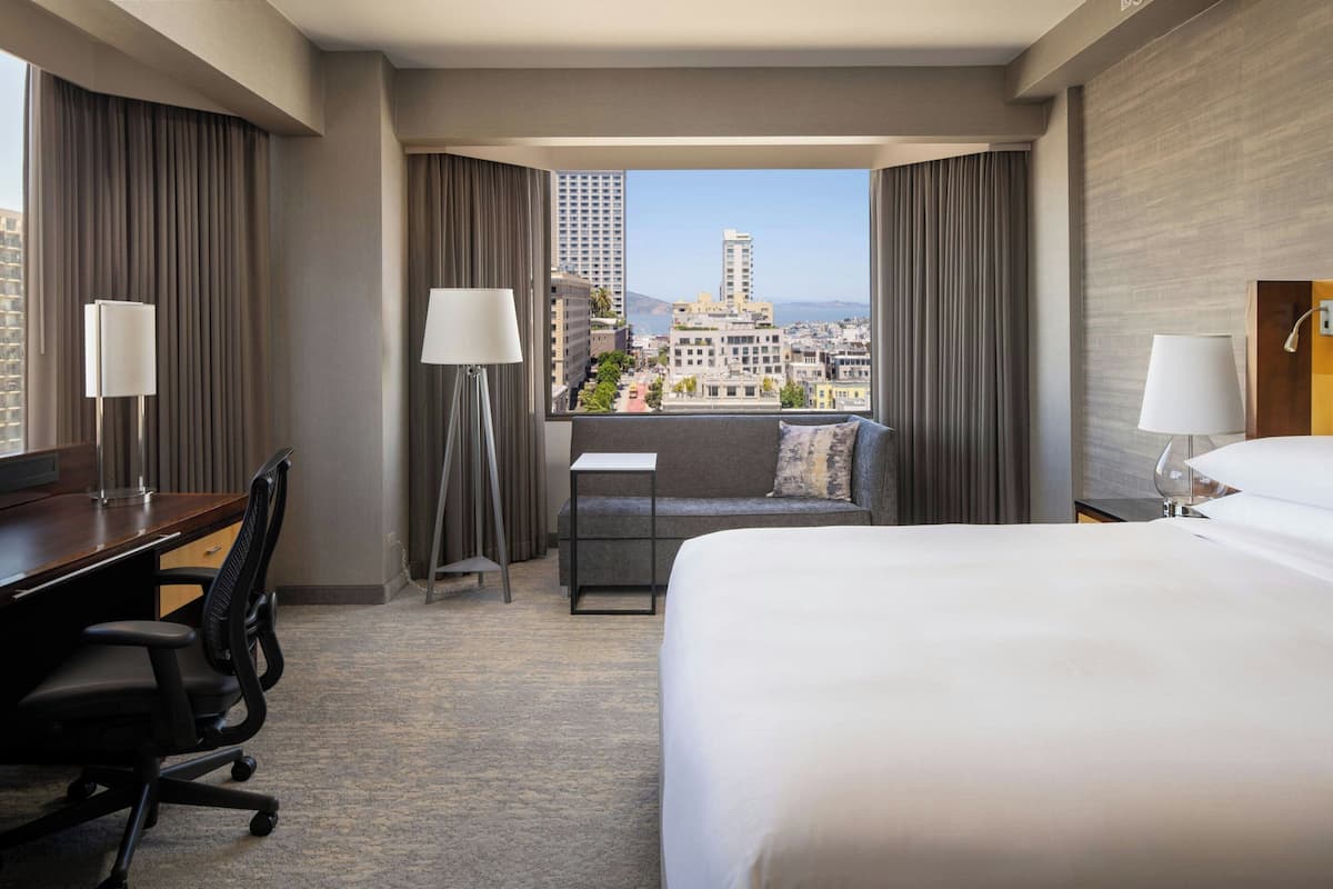 A hotel room with a large bed, a desk, a chair, and a view of the cityscape.