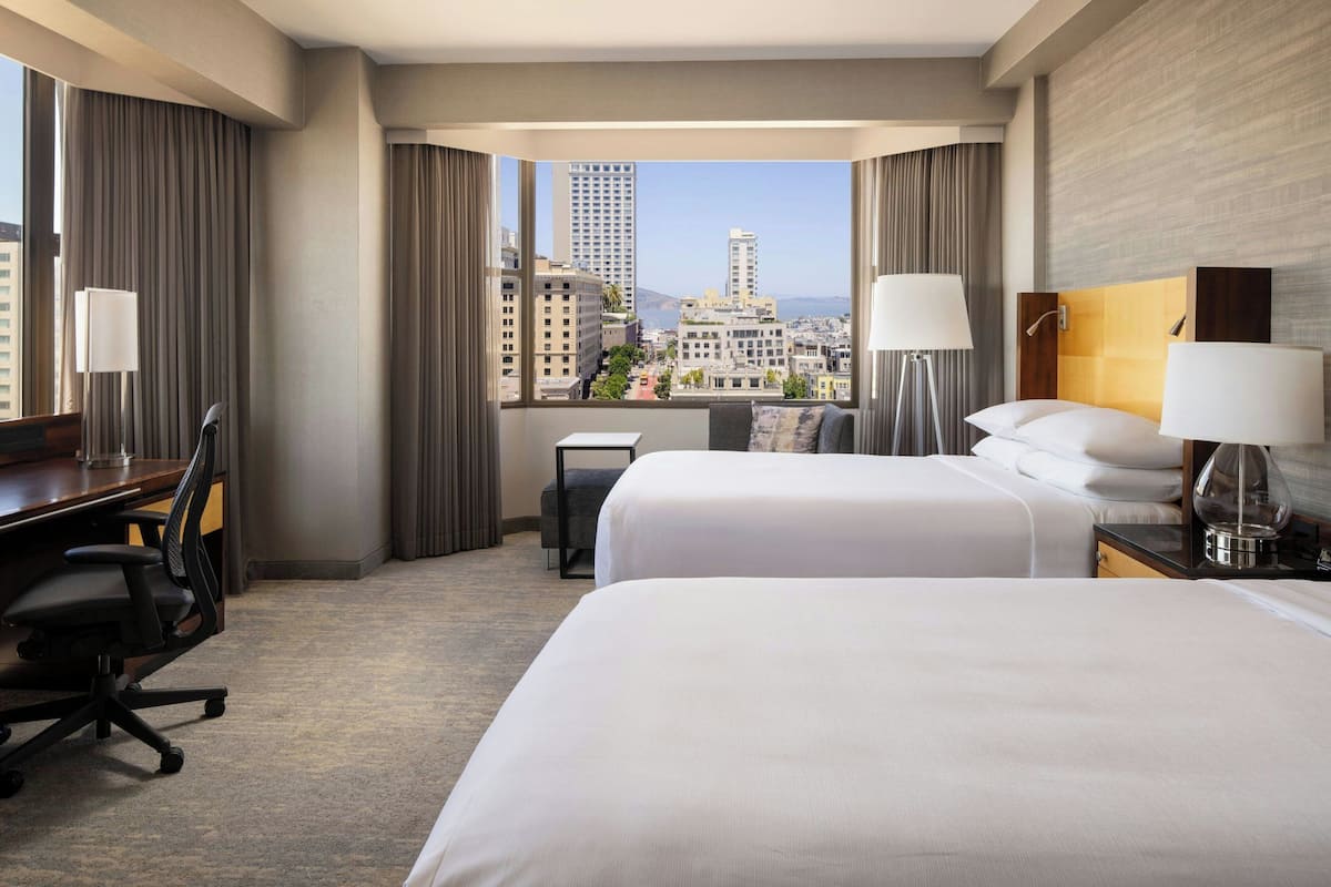 A hotel room with two beds, a desk, a chair, and a view of the cityscape.