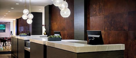 A modern reception desk with a marble top, surrounded by dark wooden panels and a hanging light fixture with spherical glass elements.