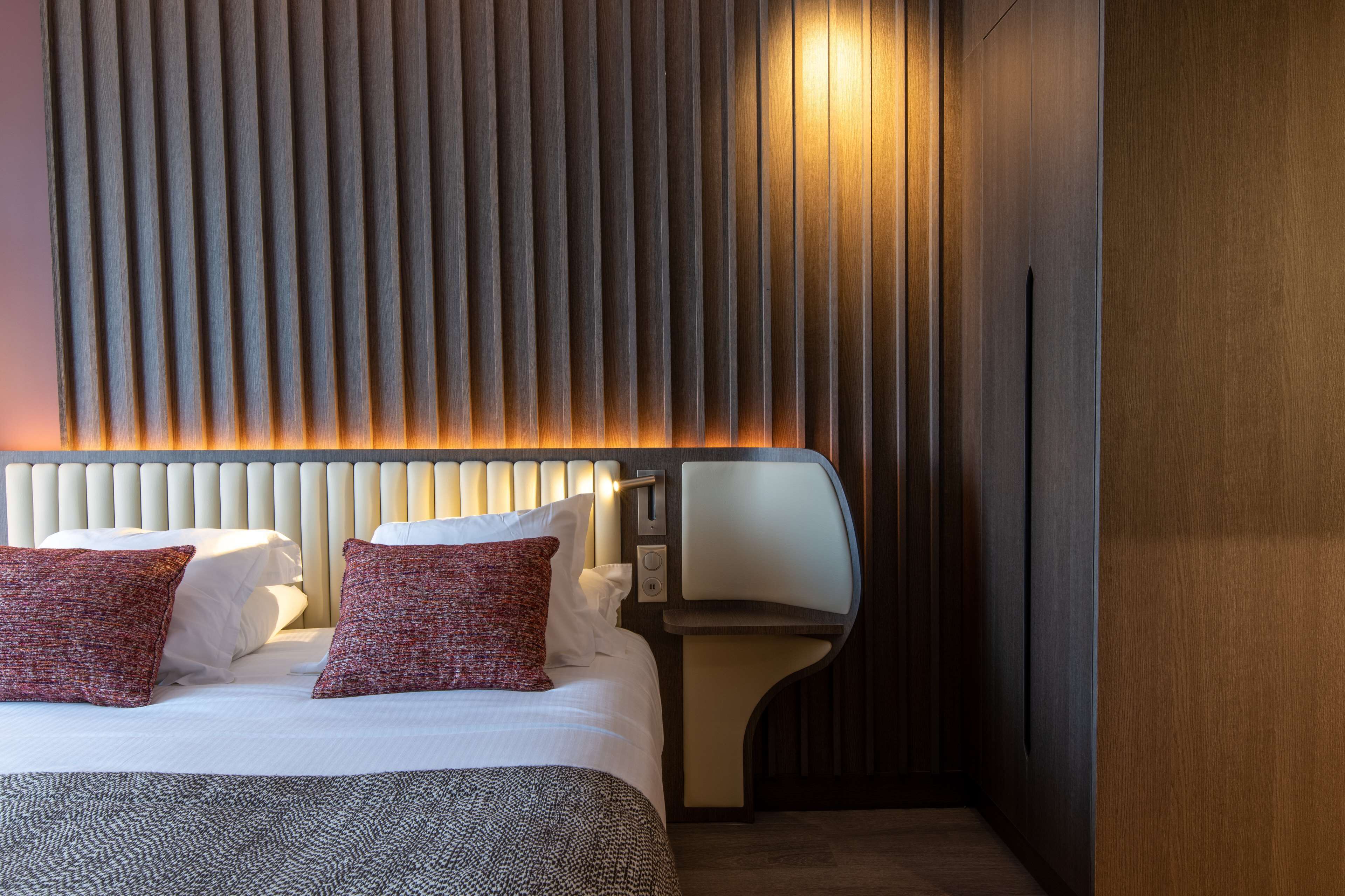 A hotel room with a bed, two pillows, a bedside table, and a wall with vertical slats.