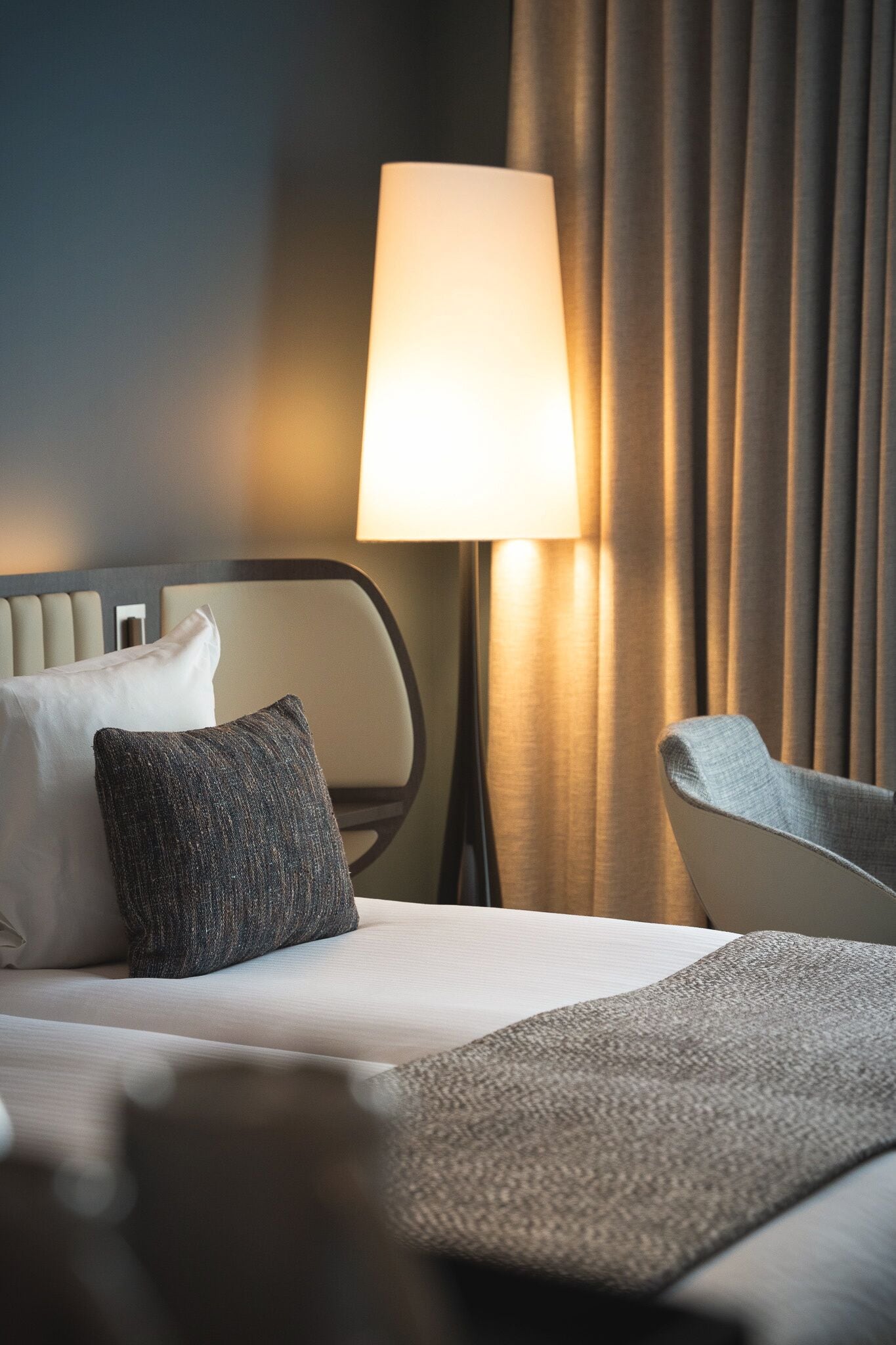 A hotel room with a bed, a chair, a lamp, and curtains.