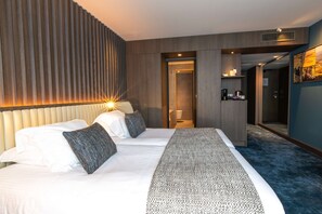 A modern hotel room with a large bed, a headboard with vertical slats, a bedside table with a lamp, and a view of the hallway.