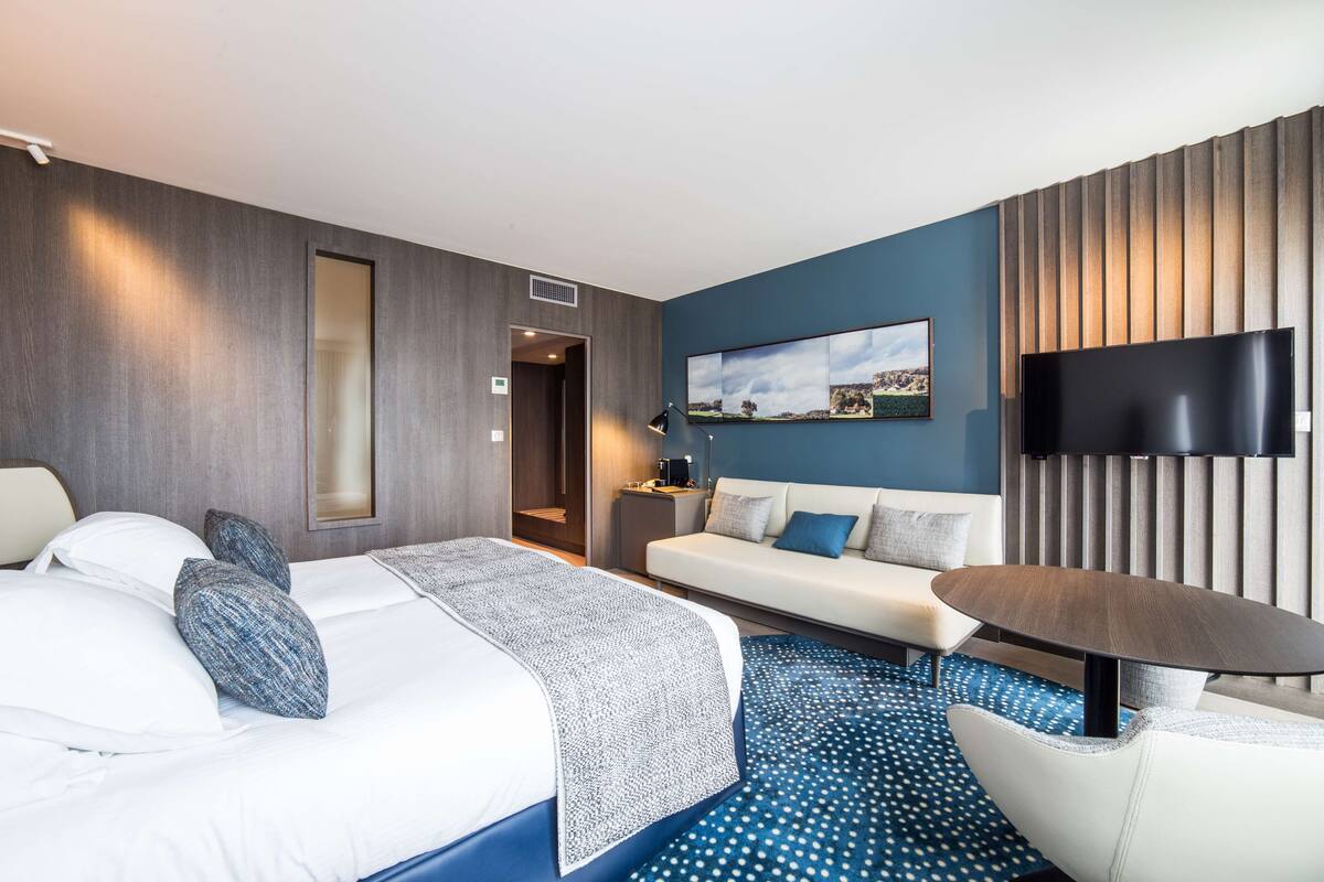 Moderni hotellihuone, jossa on sänky, sohva, televisio ja pieni pöytä.