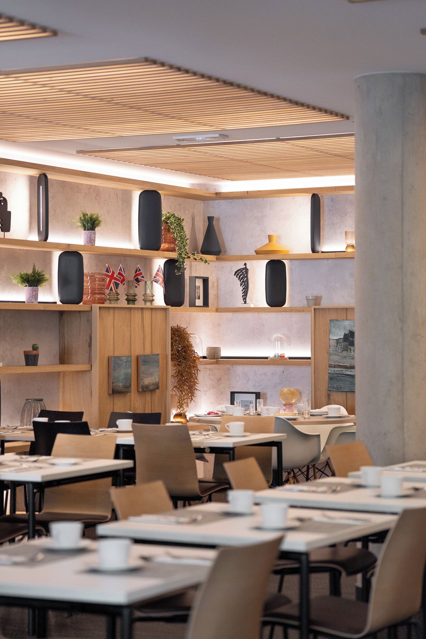 Un comedor moderno con mesas y sillas, estanterías de madera con adornos y un gran pilar.