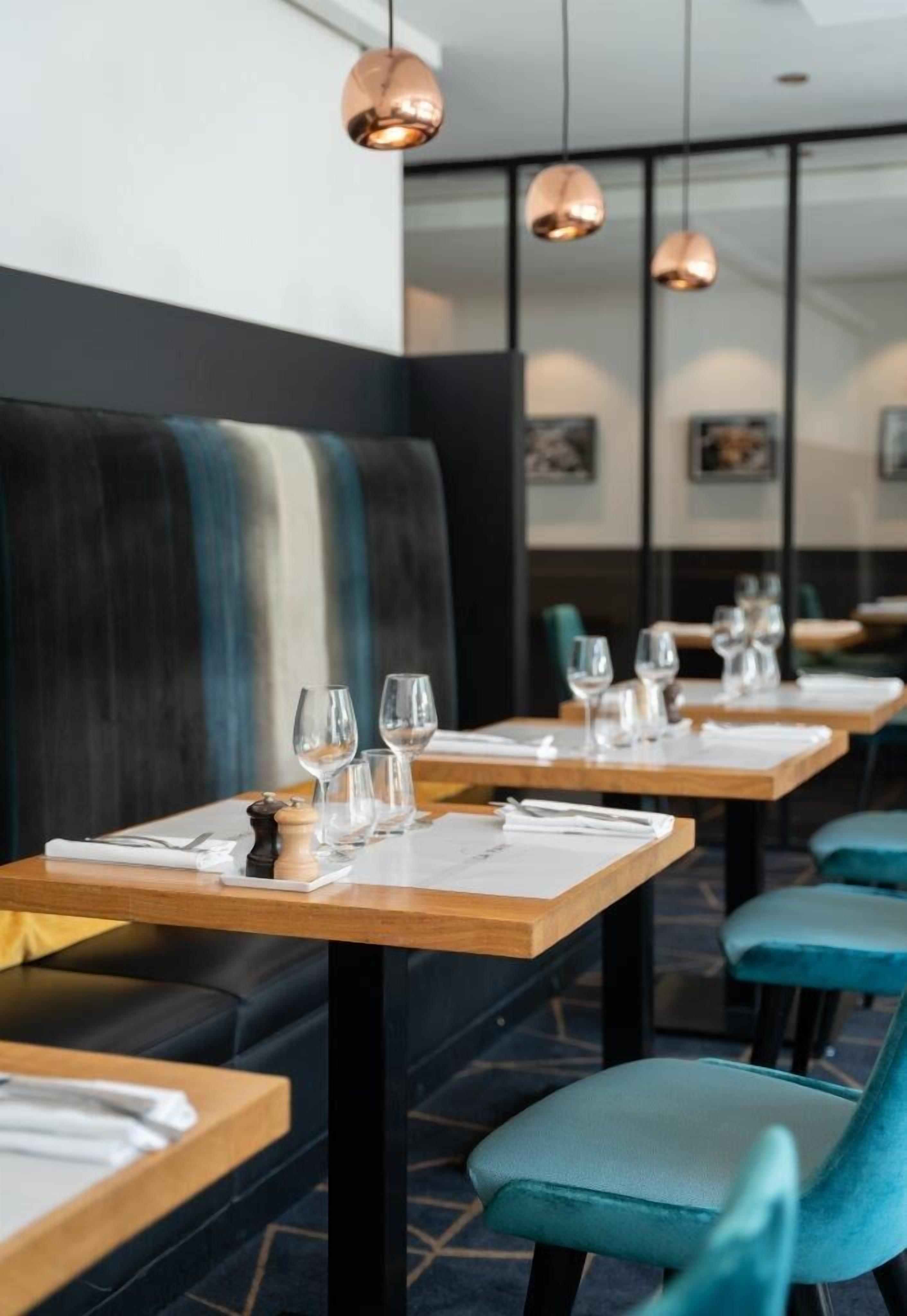 Ein Restaurantinneres mit Holztischen, blaugrünen Stühlen und Pendelleuchten aus Kupfer.