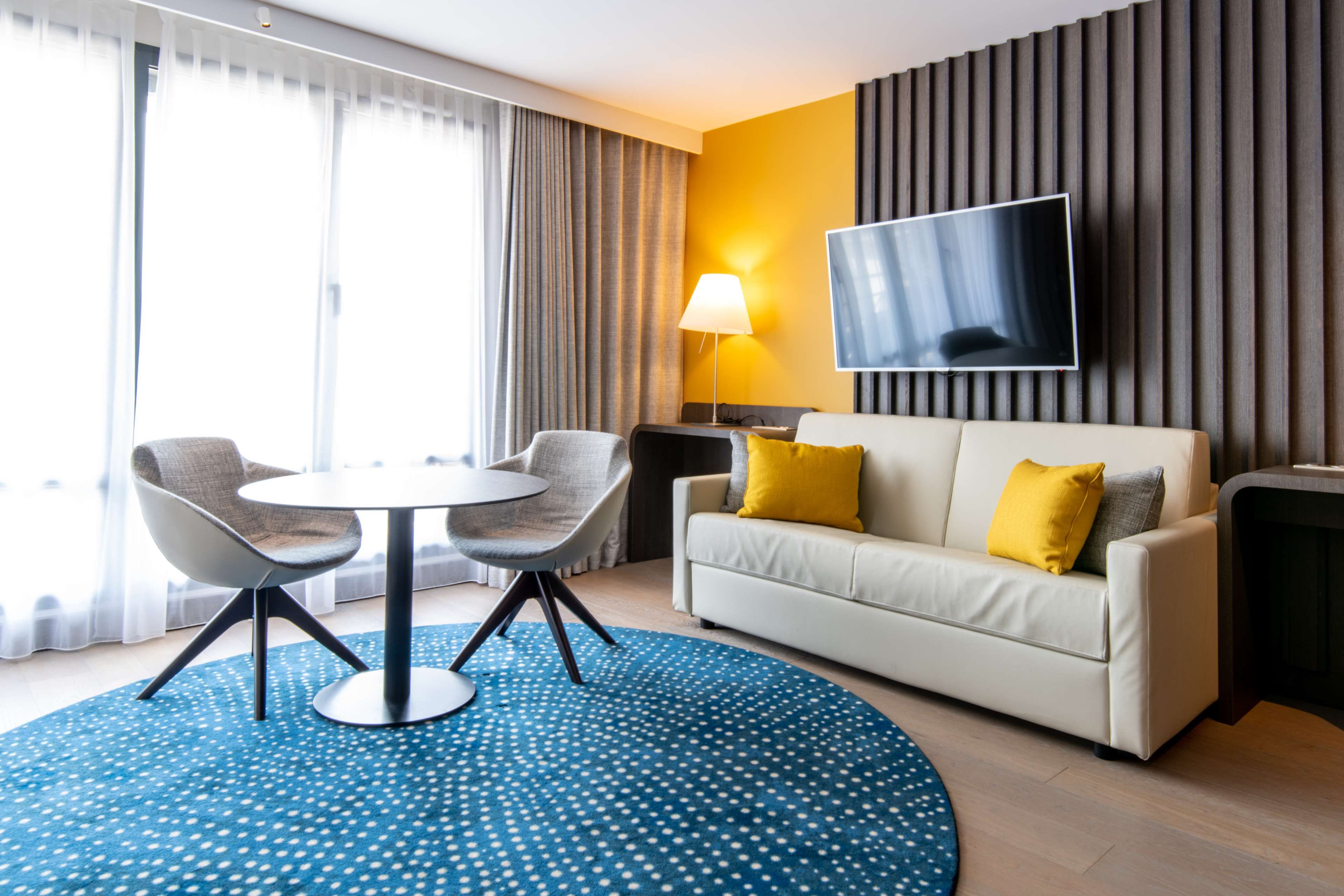 Ein modernes Hotelzimmer mit einer Couch, zwei Sesseln, einem runden Tisch, einem Flachbildfernseher und einem großen Fenster mit Vorhängen.