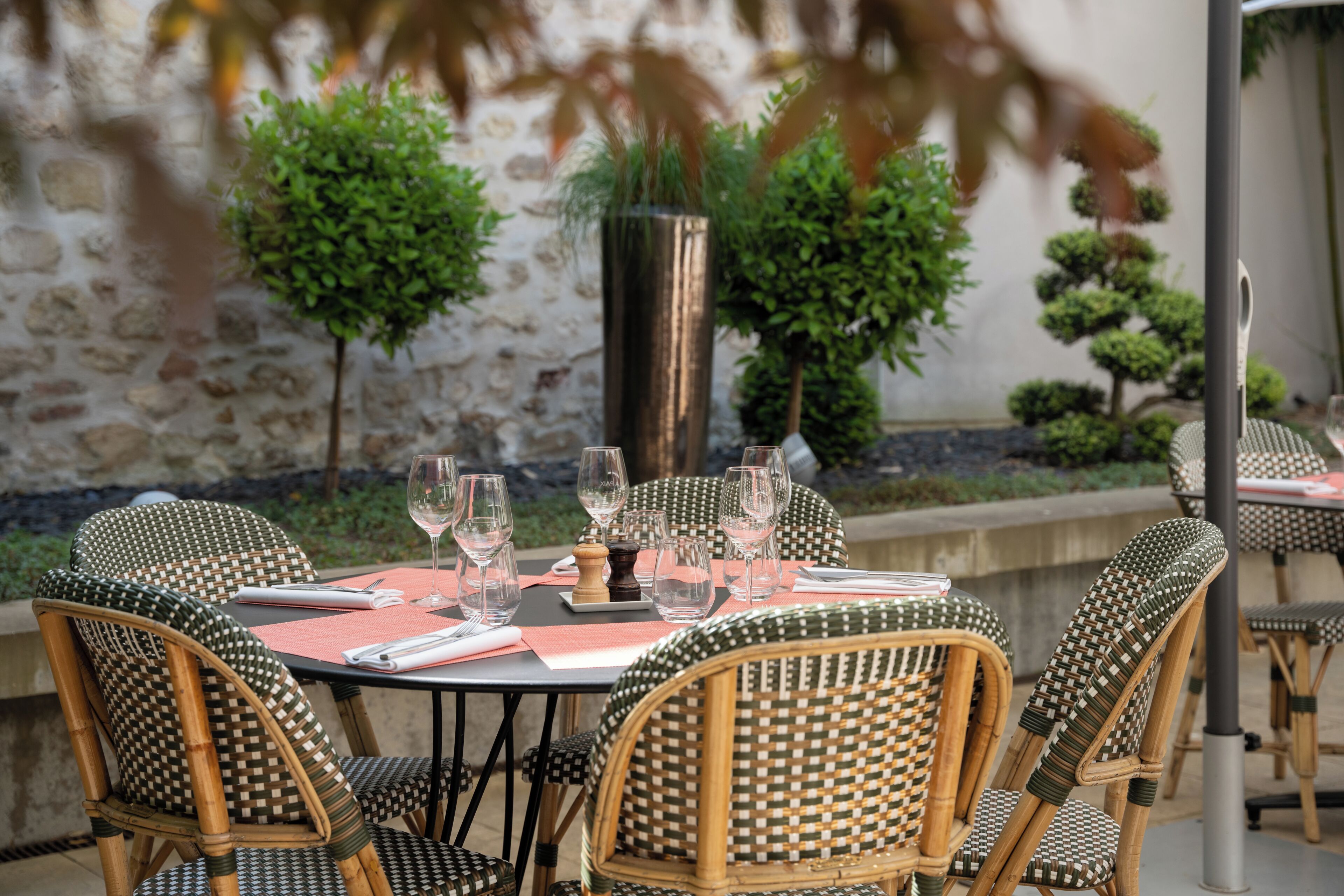 Un espace extérieur aménagé pour les repas, avec des chaises et des tables en osier, un mur en pierre et des plantes en pot.