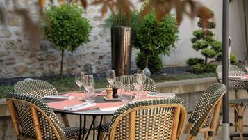 Un espace extérieur aménagé pour les repas, avec des chaises et des tables en osier, un mur en pierre et des plantes en pot.