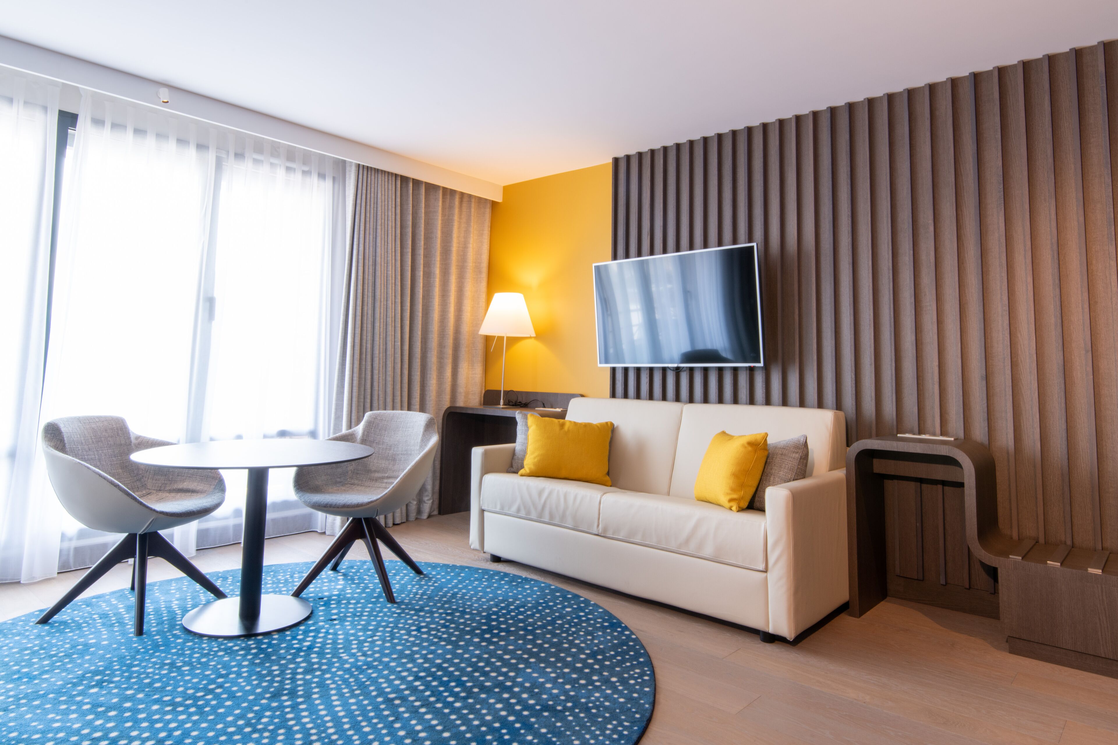 A modern hotel room with a sofa, two armchairs, a round table, a TV, and a lamp.