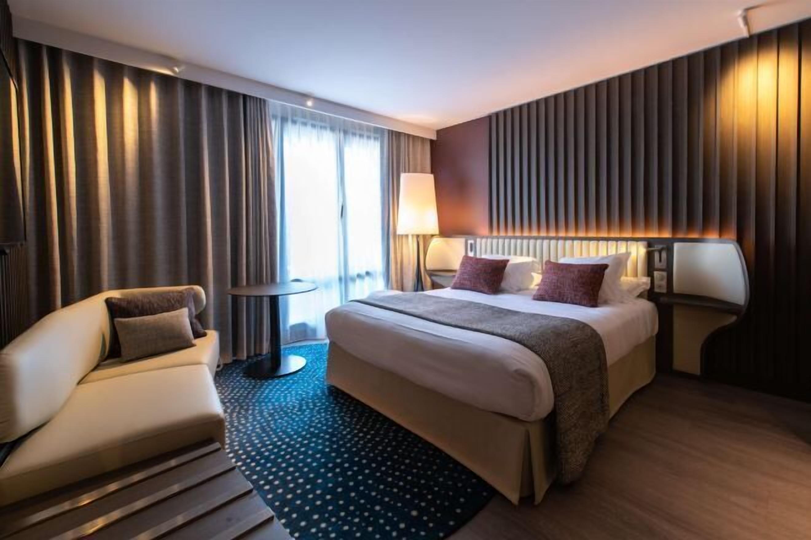 A hotel room with a bed, a sofa, a side table, a lamp, and a view of the outdoors.
