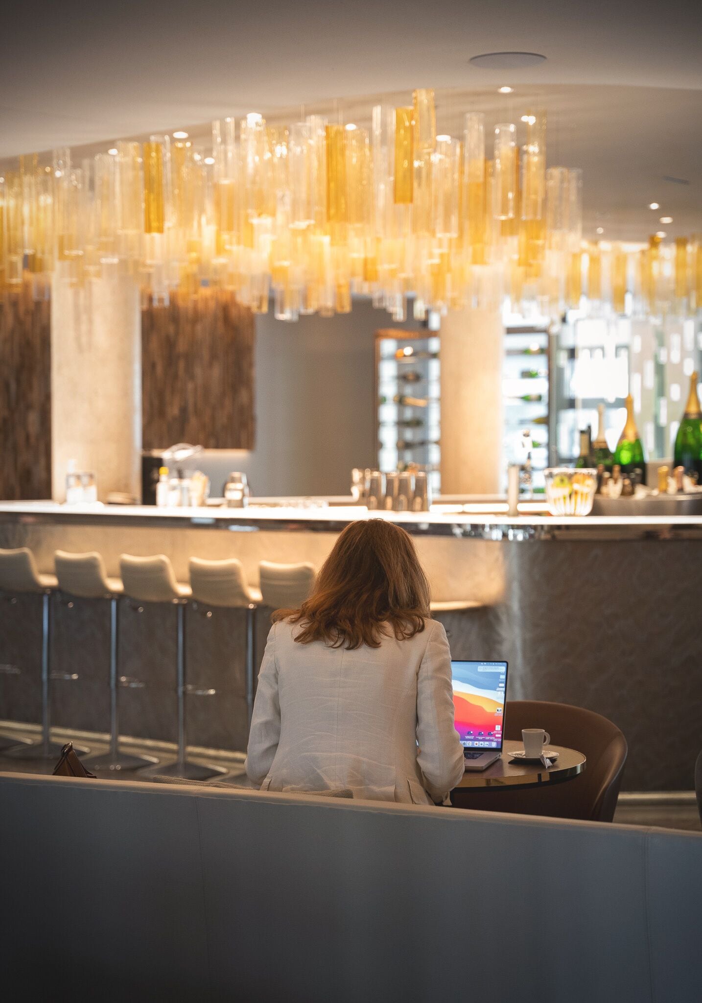 A person seated at a table with a laptop, a bar area with stools, and a modern interior design with hanging lights.