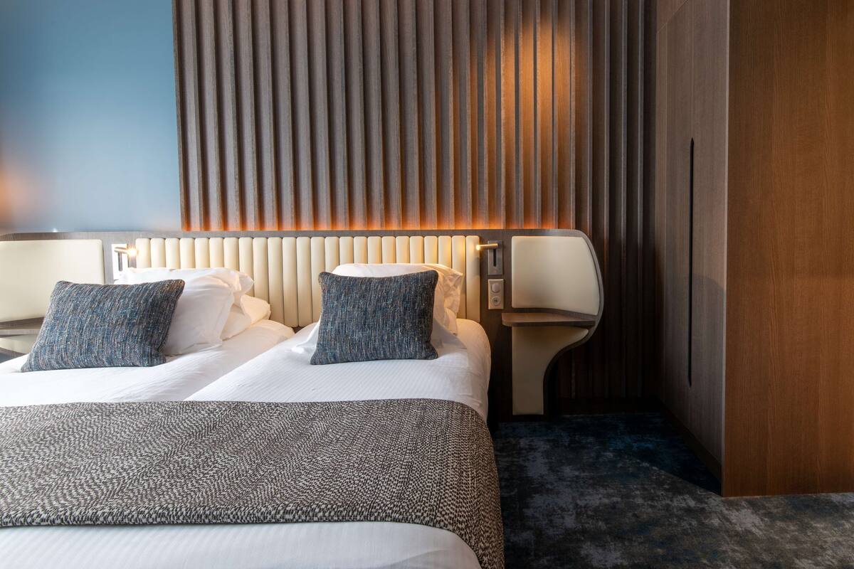Moderni hotellihuone, jossa on suuri sänky, kaksi tyynyä, yöpöytä lampun kanssa ja puupanelilla verhoiltu seinä.
