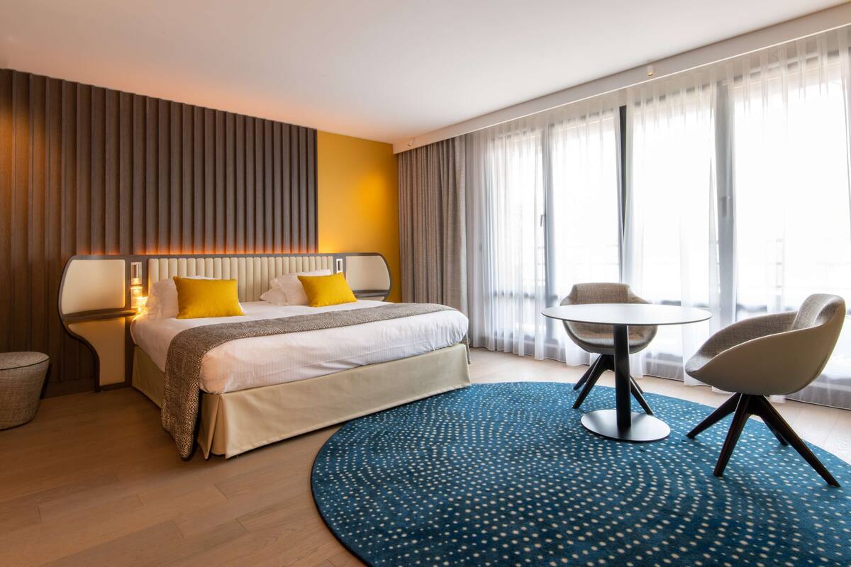 Moderni hotellihuone, jossa on suuri sänky, pyöreä ruokapöytä ja sinivärinen, kuvioitu matto.