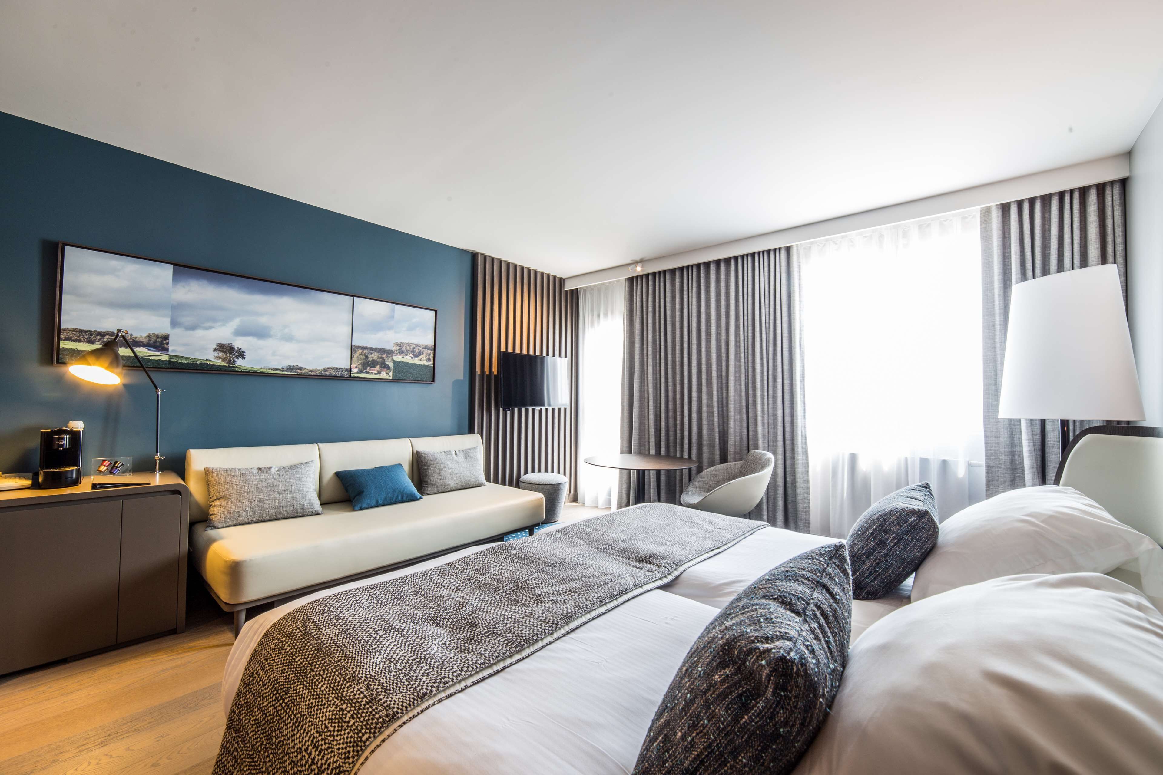 A modern hotel room with a bed, a sofa, a desk, a lamp, and a TV.