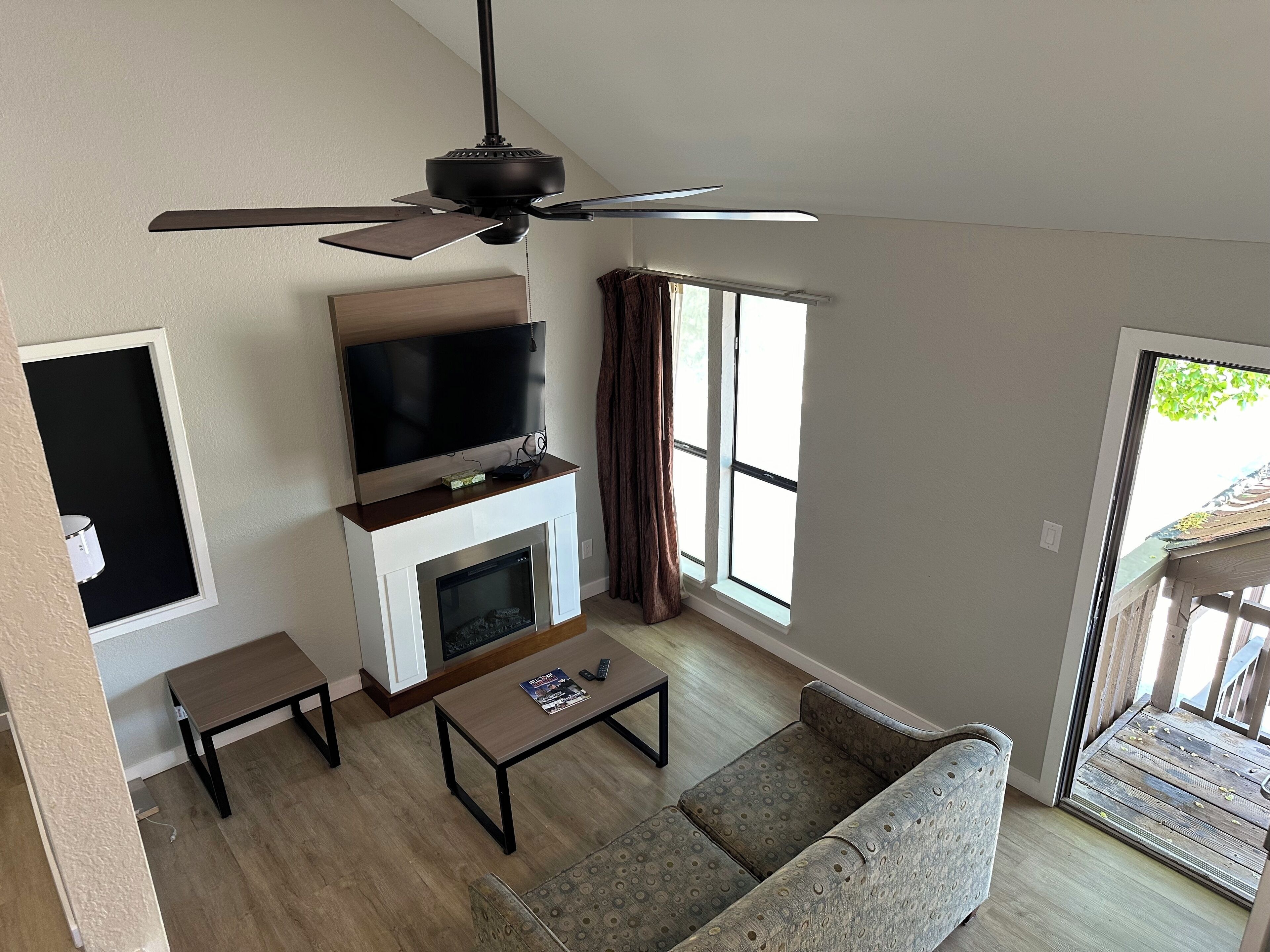 A modern living room with a flat-screen TV, fireplace, ceiling fan, and large windows.