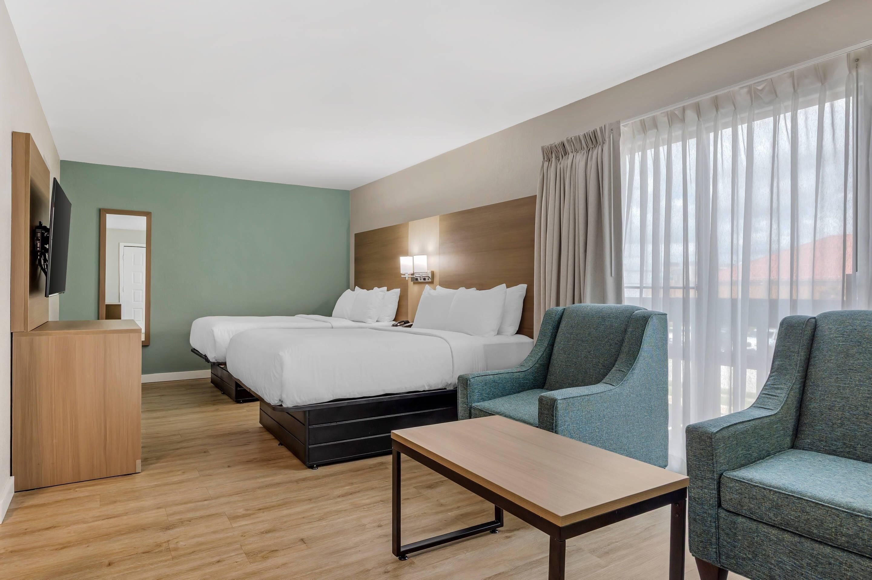 Habitación de hotel con una cama, dos sillas, una mesita, un televisor y una ventana con cortinas.