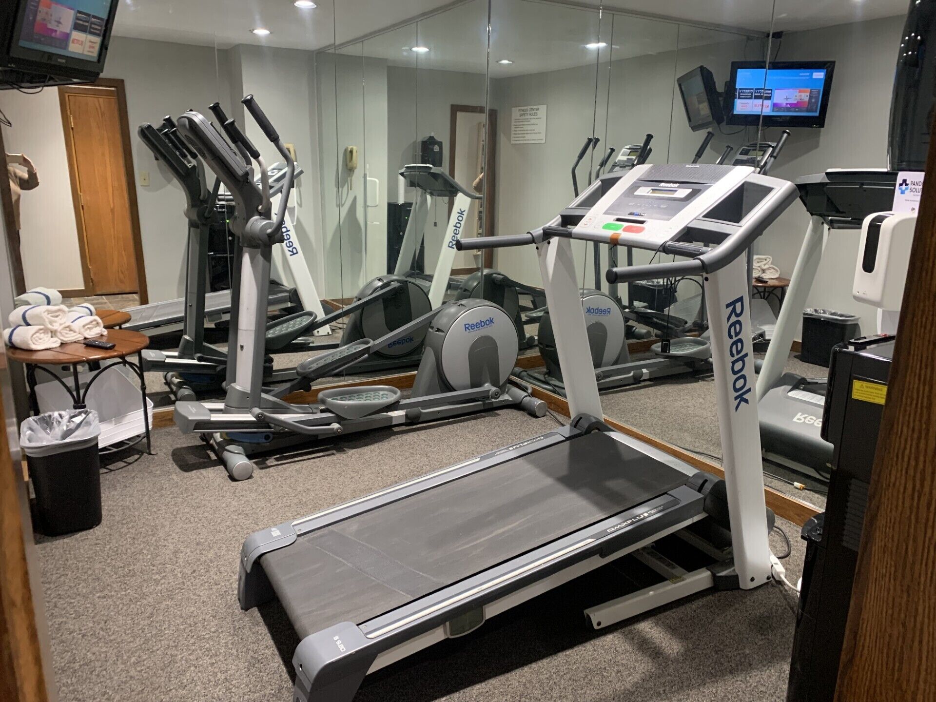 Un gimnasio con diversas máquinas de ejercicio, incluyendo cintas de correr y elípticas, y una pantalla de televisión montada en la pared.