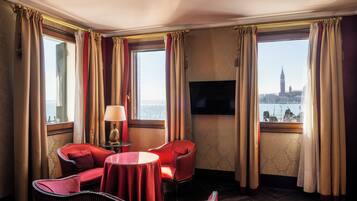 A hotel room with a large window, a flat-screen TV, a round table, and two red upholstered chairs.