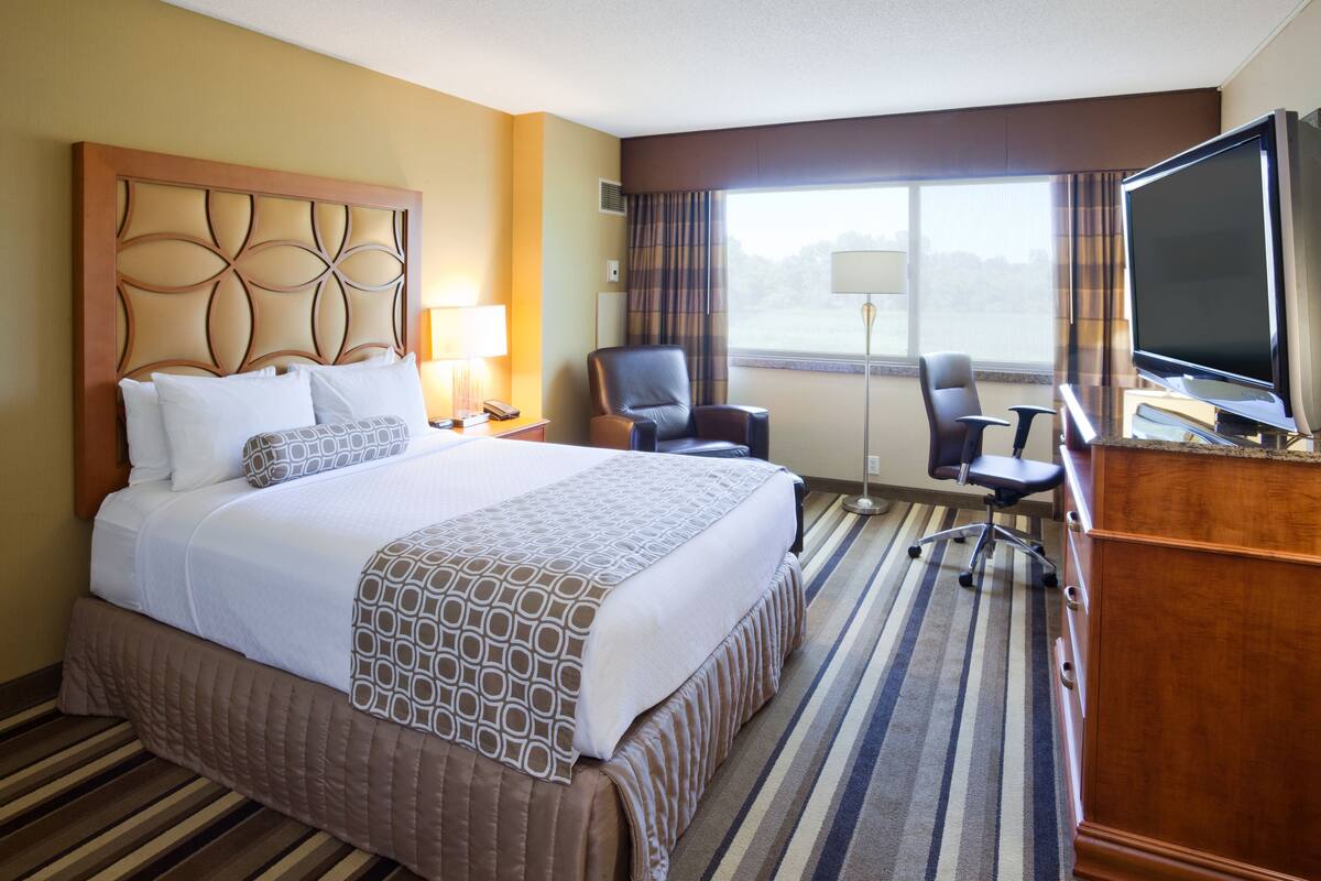 A hotel room with a large bed, a desk with a flat-screen TV, a chair, and a window with curtains.