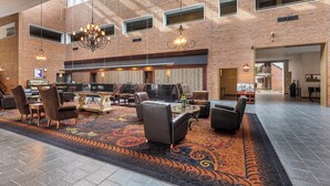 Un hall d’entrée spacieux avec un plafond haut, des murs en briques apparentes et un tapis à motifs. On y trouve plusieurs coins salon avec des fauteuils et des tables en cuir, ainsi qu’un lustre suspendu au plafond.