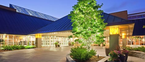 A building with a glass facade, a landscaped area with potted plants, and a tree with illuminated lights.
