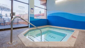 Una bañera de hidromasaje con borde de azulejos y escalera metálica, ubicada en el interior con vistas al exterior a través de amplias ventanas.