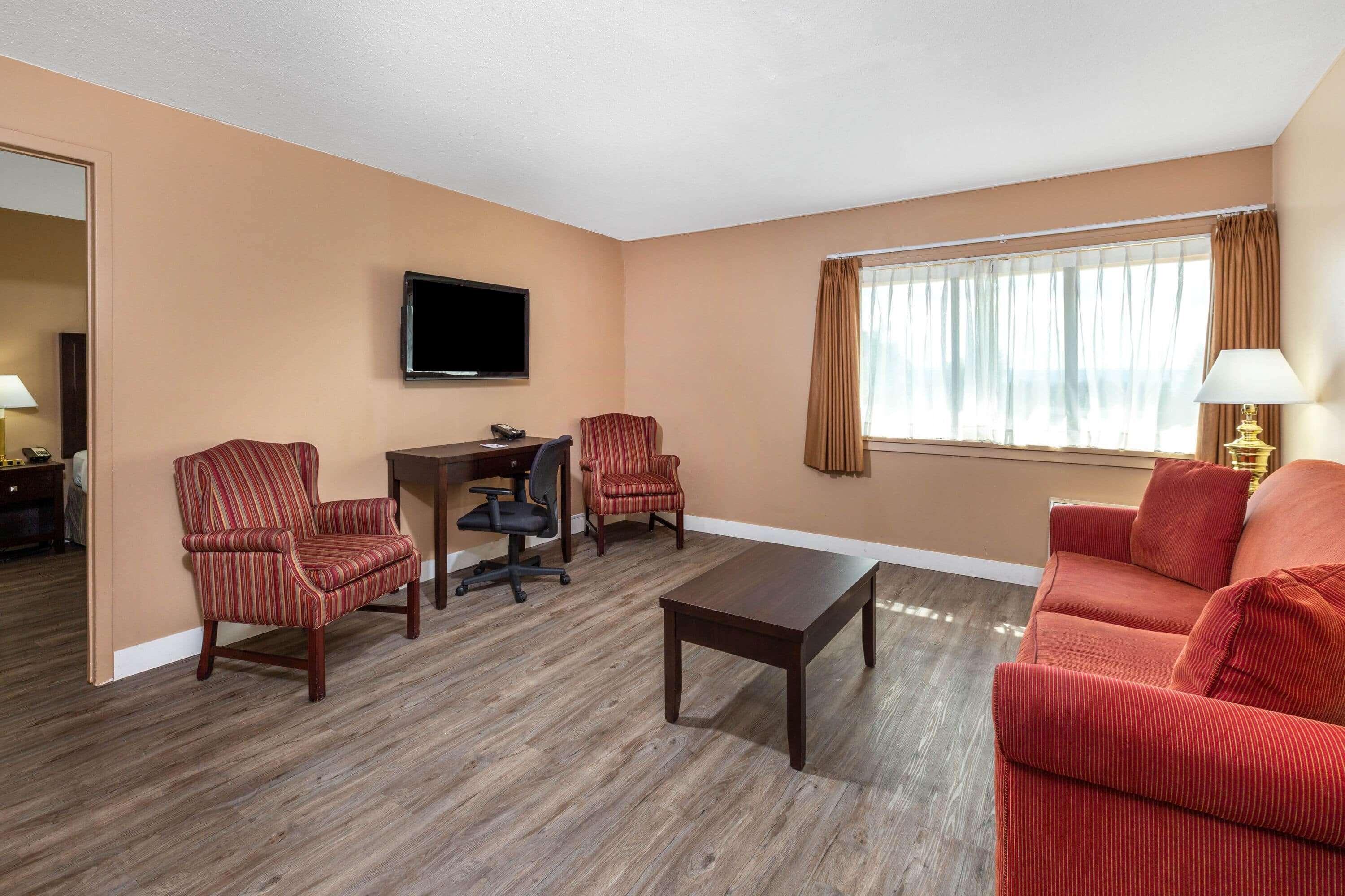 Habitación de hotel con suelo de madera, un televisor montado en la pared, un escritorio con una silla, dos sillones, un sofá y una ventana con cortinas.