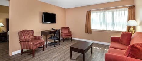 A hotel room with a wooden floor, a TV mounted on the wall, a desk with a chair, two armchairs, a sofa, and a window with curtains.