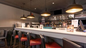 Zona de bar con una barra larga, varios taburetes, luces colgantes y estantes llenos de botellas y vasos.