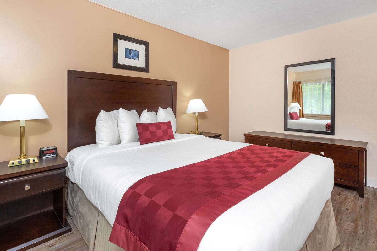 Ein Hotelzimmer mit einem großen Bett, zwei Nachttischen mit Lampen, einem Schminktisch und einem Spiegel. An der Wand hängt ein gerahmtes Bild.