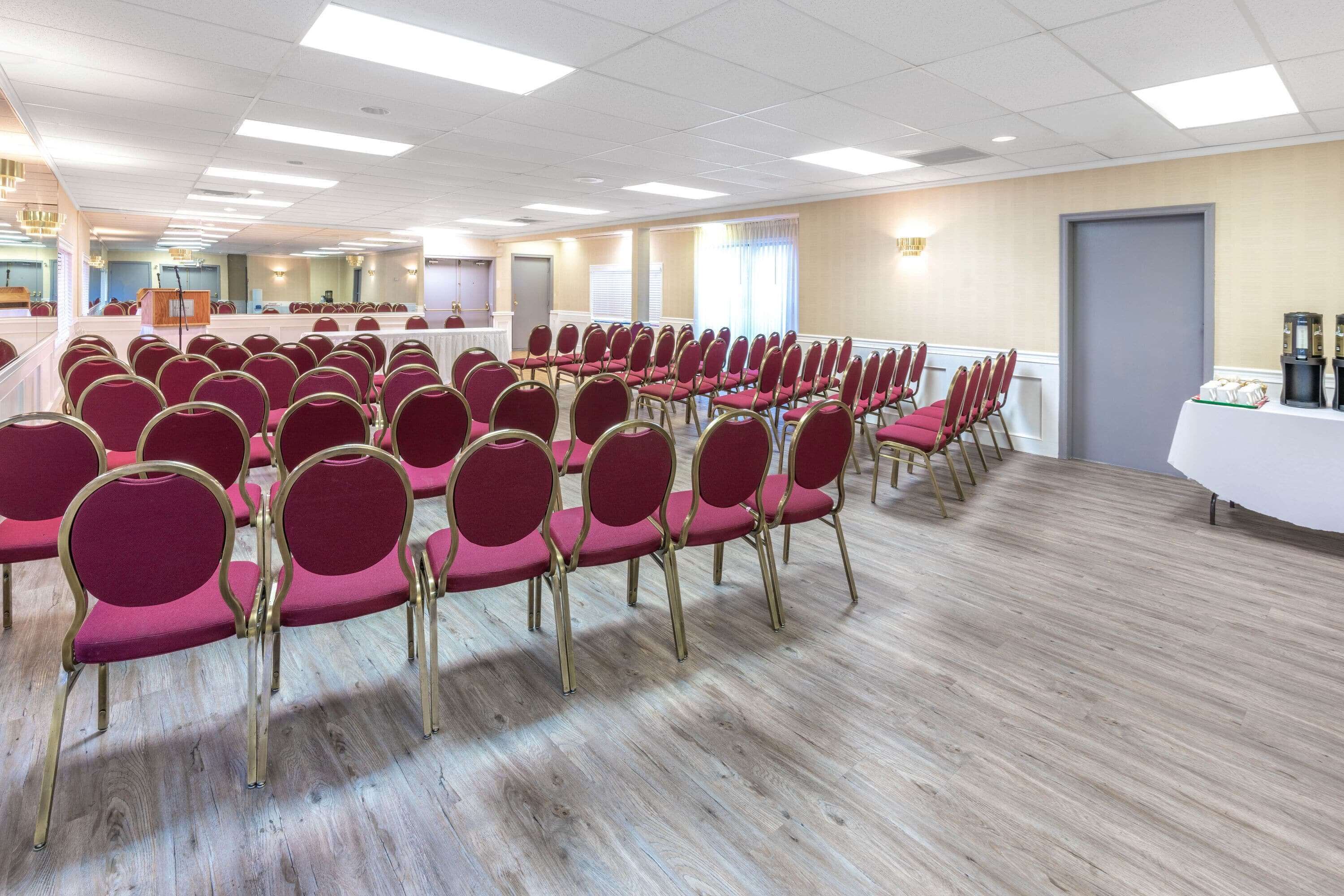 A conference room with rows of red chairs and a white table, wooden flooring, and ceiling lights.