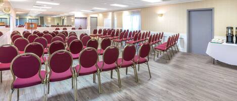 A conference room with rows of red chairs and a white table, wooden flooring, and ceiling lights.